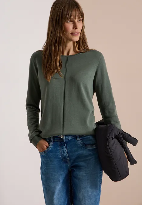 Frau in grünem Pullover, Jeans und hält eine dunkle Jacke in der Hand.