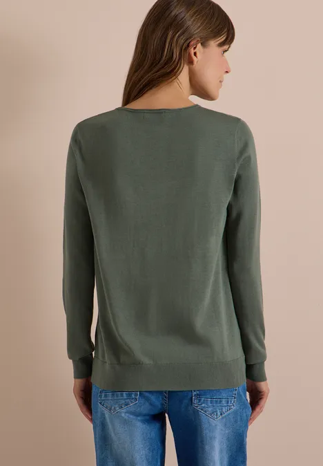 Eine Frau von hinten, trägt einen schlichten grünen Pullover und blaue Jeans vor neutralem Hintergrund.