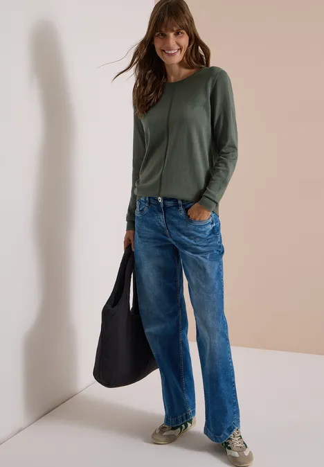 Frau in legerem Outfit: grünes Langarmshirt, lockere Jeans, Sneaker und schwarze Tasche vor weißer Wand.