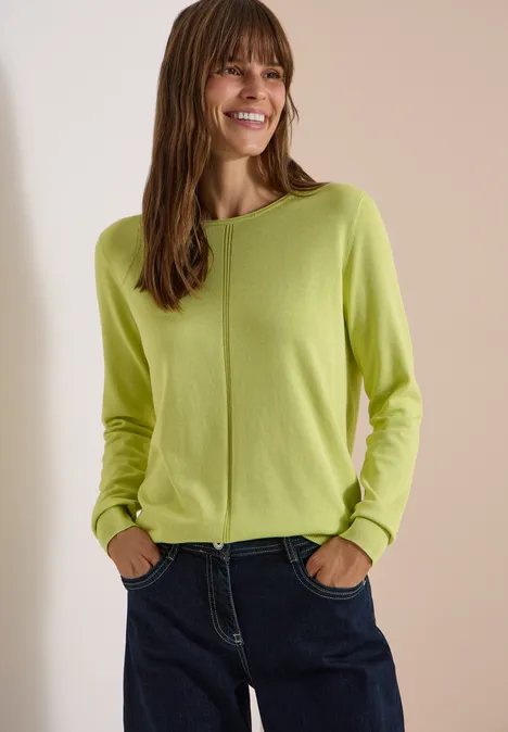 Eine lächelnde Frau trägt einen hellgrünen Pullover und dunkle Jeans vor neutralem Hintergrund.