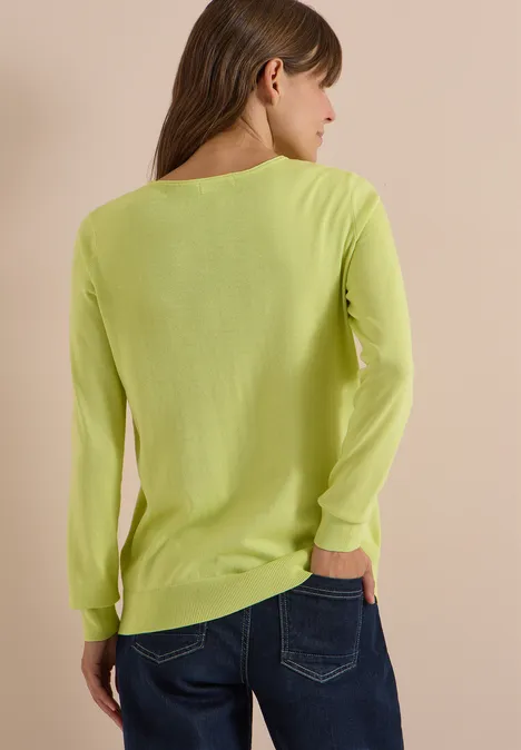 Frau von hinten in neongelbgrünem Langarmshirt und blauen Jeans vor beigem Hintergrund.