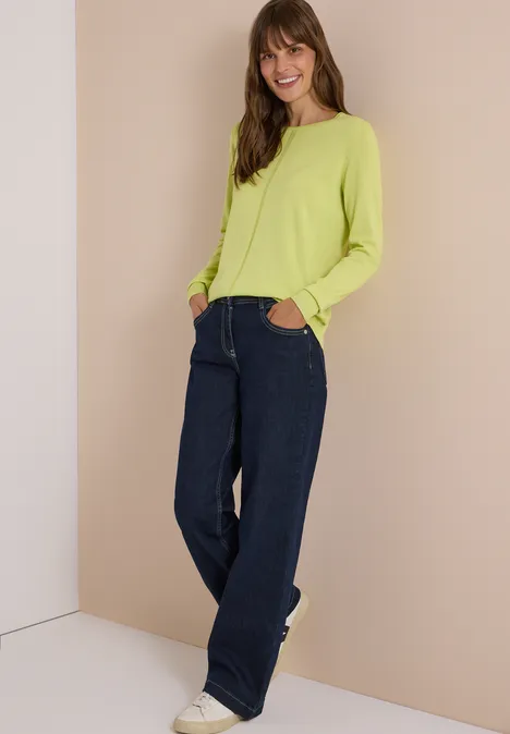 Frau in limettengrünem Pullover, weiten Jeans und weißen Sneakern lehnt lächelnd entspannt an der Wand.