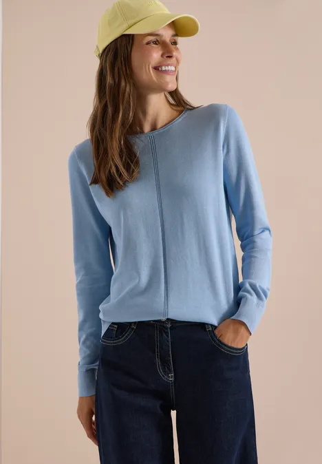 Frau in hellblauem Pullover, Jeans und gelbgrüner Kappe vor neutralem Hintergrund, lächelnd seitlich schauend.