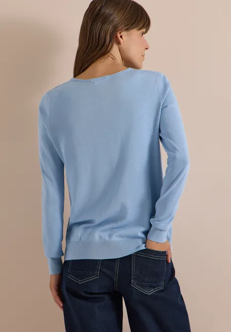 Frau von hinten in hellblauem Langarm-Pullover und Jeans vor beigem Hintergrund.