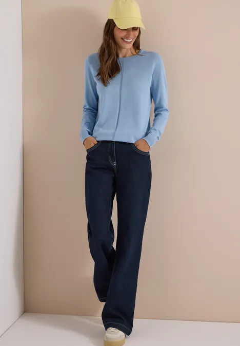 Eine lächelnde Frau trägt eine gelbe Kappe, einen hellblauen Pullover, weite dunkle Jeans und weiße Sneaker.