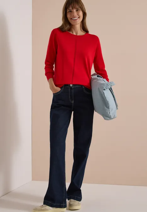 Femme souriante en pull rouge et jean évasé bleu foncé, tenant un sac bleu clair.