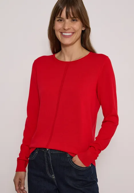 Une femme souriante porte un pull rouge et un jean sur fond clair.