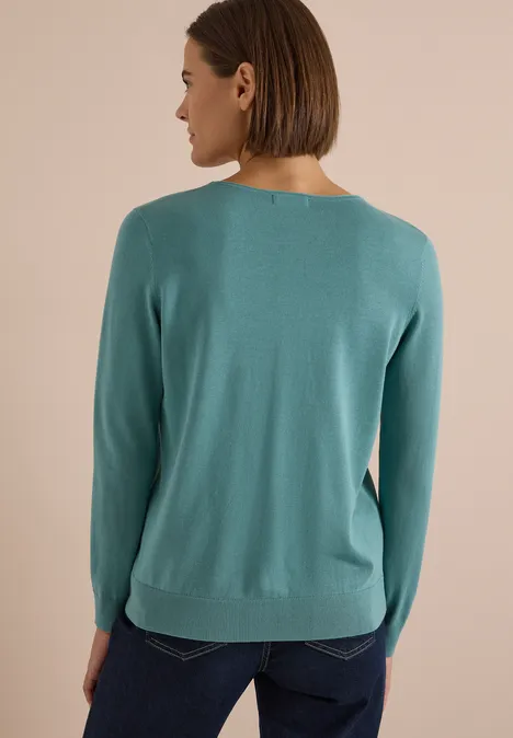 Eine Frau von hinten in einem türkisfarbenen Pullover und Jeans vor neutralem Hintergrund.