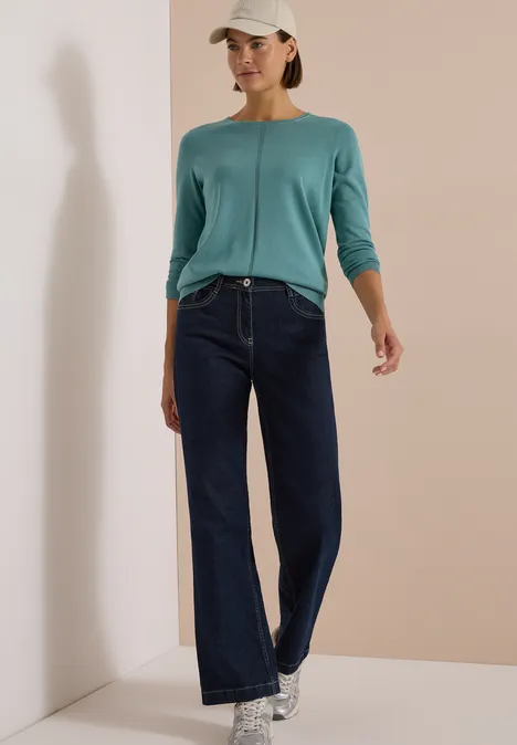 Frau in blaugrünem Langarmshirt, weit geschnittenen Jeans, Sneakern und Cap vor beige Wand.