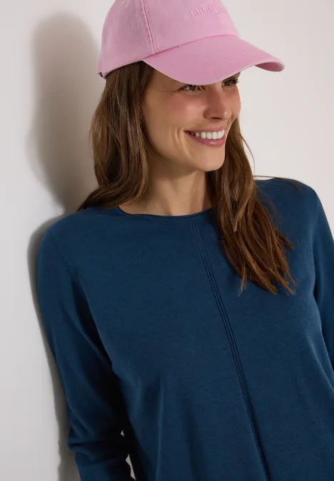 Basic Pullover mit Rundhals in Unifarbe moonlight blue Eine lächelnde Frau mit rosa Cap und blauem Oberteil vor heller Wand.