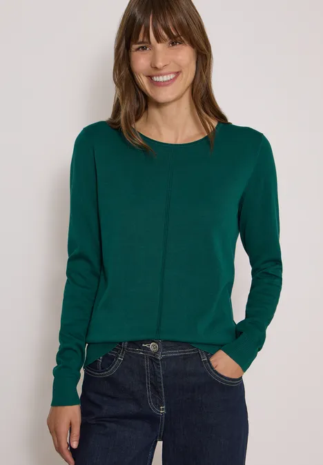 Jeune femme souriante en pull vert et jean devant un fond neutre.