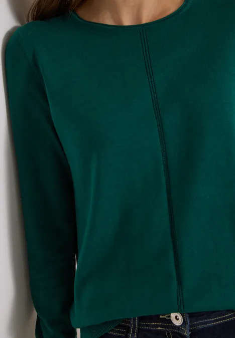 Gros plan sur un pull vert foncé à surpiqûre centrale porté avec un jean bleu.