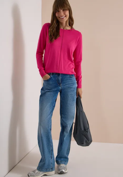 Basic Pullover mit Rundhals in Unifarbe pink flash Frau in pinkem Pullover und weitem Jeans mit grauer Tasche und weißen Sneakern lehnt lässig an Wand.