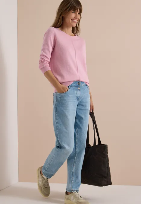 Femme en pull rose et jean, portant des baskets vertes et un grand sac noir, debout devant un mur clair.