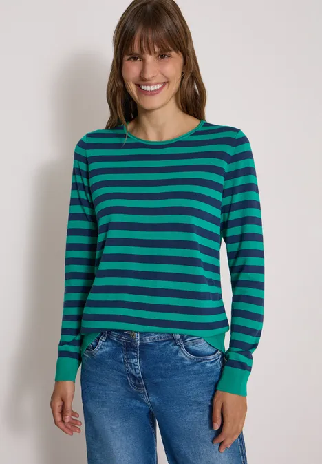 Frau in grün-blau gestreiftem Pullover und Jeans vor neutralem Hintergrund.