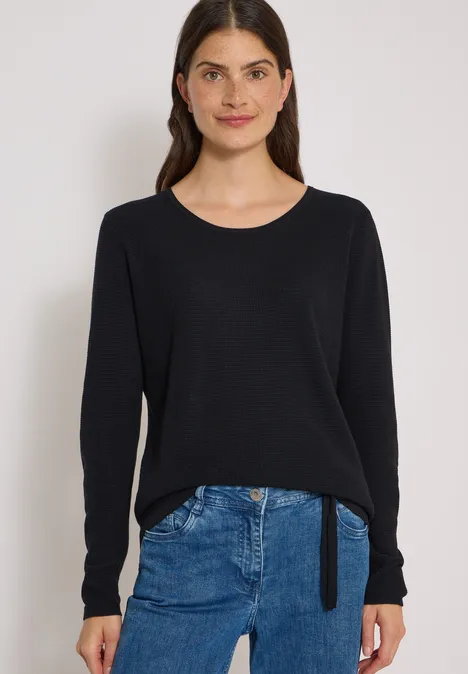 Eine Frau trägt einen schwarzen Pullover und eine blaue Jeans vor hellem Hintergrund.