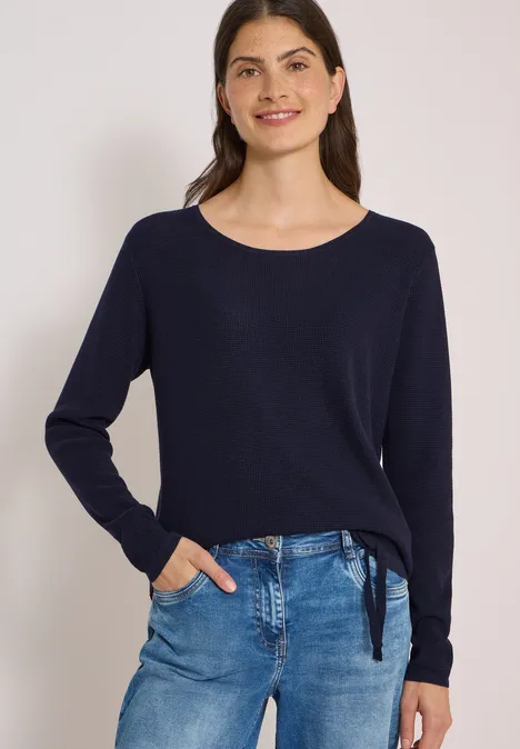 Frau in dunkelblauem Pullover und Jeans, lächelnd, vor hellem Hintergrund.