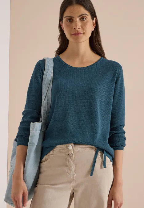 Pull à col rond et texturé dull teal blue melange Frau trägt blauen Pullover, helle Hose und Jeans-Tasche vor neutralem Hintergrund.