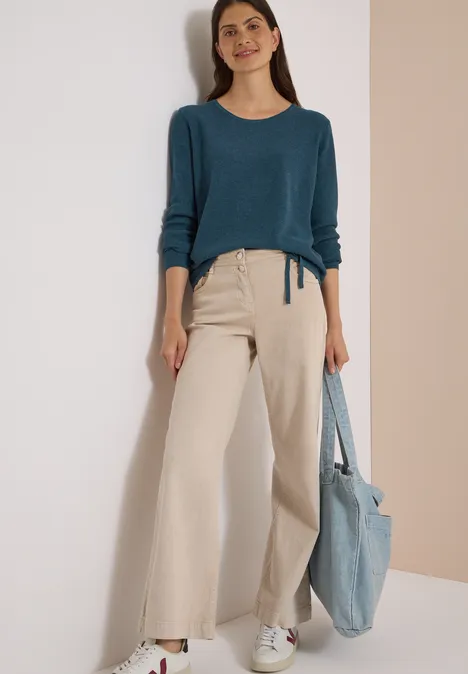 Pull à col rond et texturé dull teal blue melange Lächelnde Frau mit blauem Pullover, beigen Culottes, weißen Sneakern und hellblauer Tasche vor einer Wand.