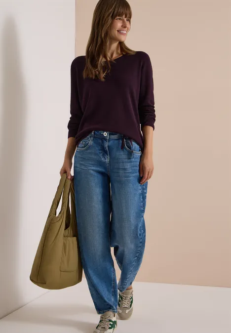 Frau in lila Pullover, weiten Jeans und Sneakers, trägt eine große grüne Tasche und lächelt entspannt.