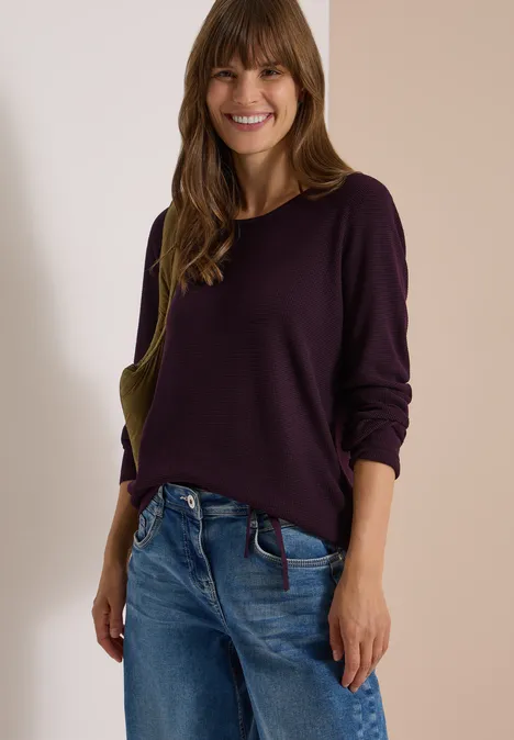 Frau mit lila Pullover und Jeans lächelt vor neutralem Hintergrund.