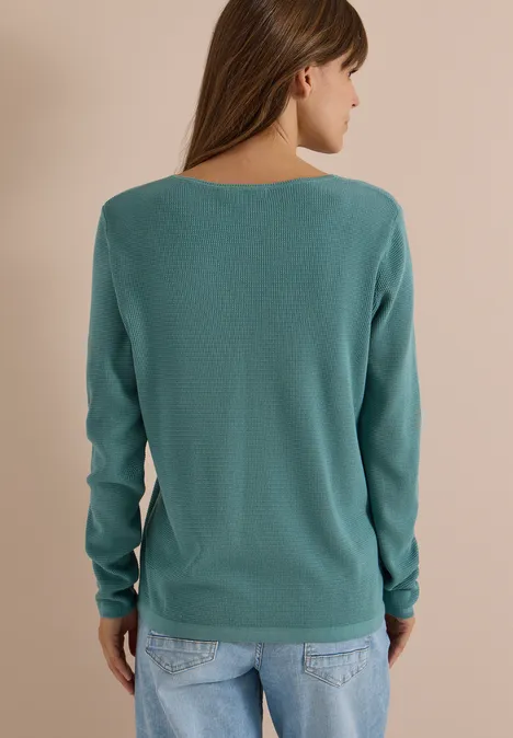 Une femme de dos porte un pull turquoise et un jean devant un fond uni.