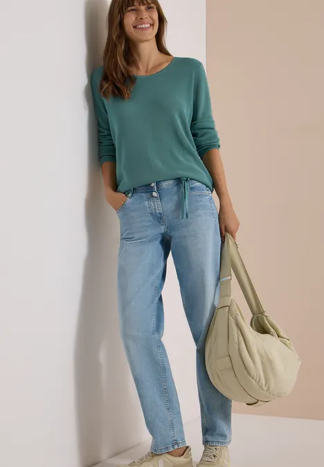 Une femme appuyée contre un mur porte un haut vert, un jean bleu et un grand sac en bandoulière.