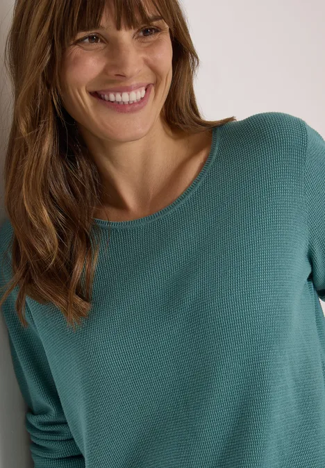 Femme souriante en pull turquoise, portrait mi-corps sur fond clair.