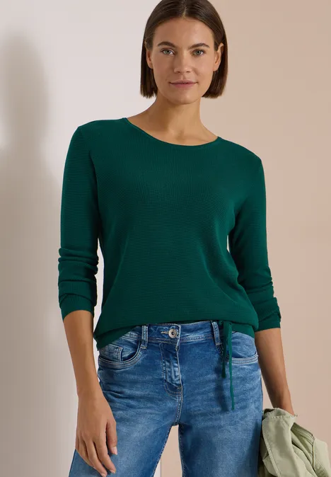 Frau mit dunkelgrünem Pullover und blauen Jeans steht vor neutralem Hintergrund und hält eine Jacke.