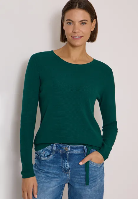 Junge Frau mit dunkelgrünem Pullover und Jeans lächelt entspannt mit einer Hand in der Hosentasche.