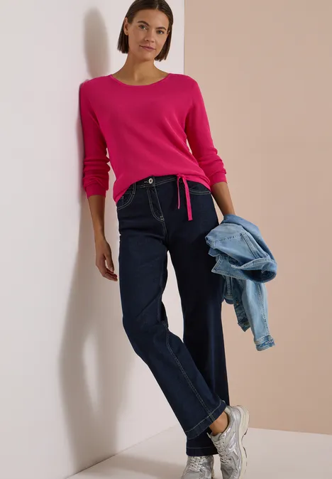 Frau lehnt an Wand in pinkem Pullover, dunklen Jeans, weißen Sneakers und hält eine Jeansjacke in der Hand.