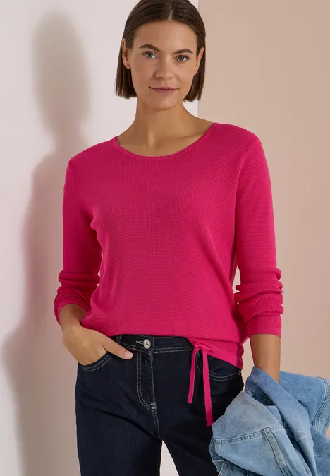 Frau in pinkem Pullover, dunkler Jeans, mit Jeansjacke vor beiger Wand.