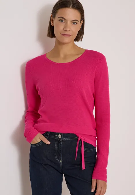 Frau in pinkem Rundhals-Pullover mit seitlichem Bindeband und dunkler Jeans vor hellem Hintergrund.