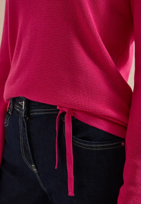 Pinkfarbener Pullover mit seitlicher Schlaufenbindung und langen Ärmeln, kombiniert mit dunkler Jeans.