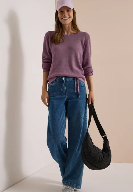 Femme souriante en pull violet, casquette assortie, jean large et sac noir à l'épaule, adossée à un mur.