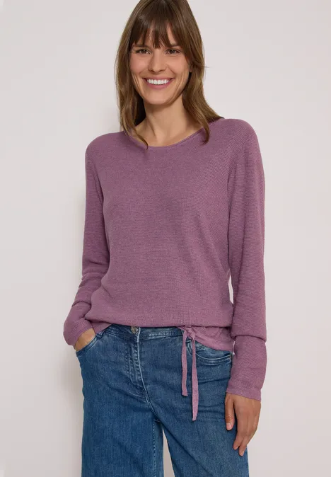 Une femme souriante porte un pull violet et un jean bleu devant un fond clair.