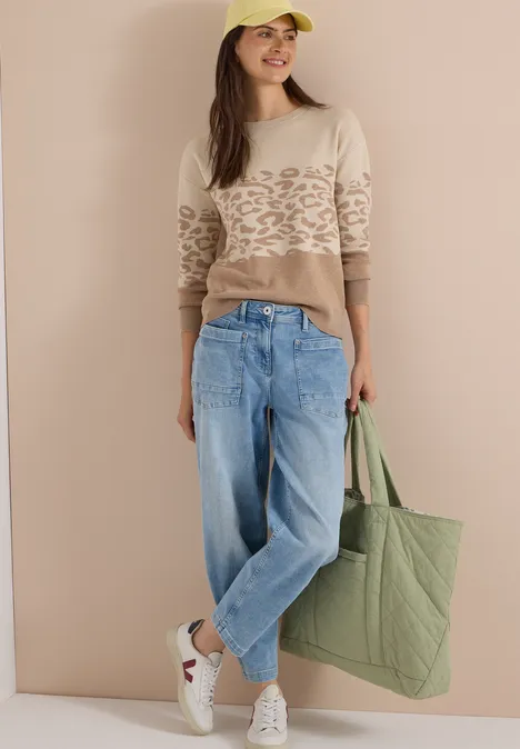 Langarm Pullover mit Leo-Jacquard-Muster creamy mocha melange Model trägt beigefarbenen Pullover mit Muster, weite Jeans, weiße Sneaker und grüne Stofftasche.