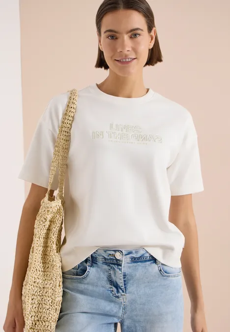 Bluza z krótkim rękawem z haftem vanilla white Kobieta w białej koszulce, dżinsach i plecionej torbie na neutralnym tle.