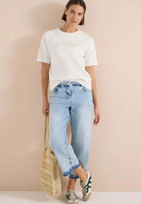 Bluza z krótkim rękawem z haftem vanilla white Kobieta w białym T-shircie, jasnych dżinsach, zielono-białych butach, trzyma słomianą torebkę na beżowym tle.