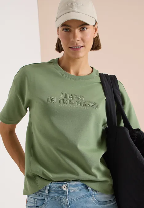 Bluza z krótkim rękawem i haftem deep matcha green Osoba w jasnooliwkowej koszulce, dżinsach, beżowej czapce i czarnej torbie na neutralnym tle