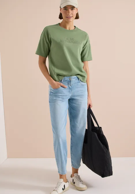 Bluza z krótkim rękawem i haftem deep matcha green Kobieta ma zieloną koszulkę, jasne dżinsy i trzyma czarną torbę