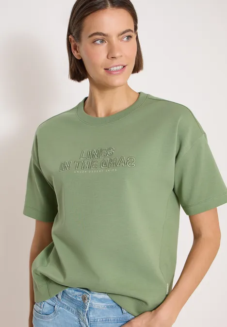 Bluza z krótkim rękawem i haftem deep matcha green Osoba nosi oliwkowy t-shirt z nadrukiem i jasnoniebieskie dżinsy
