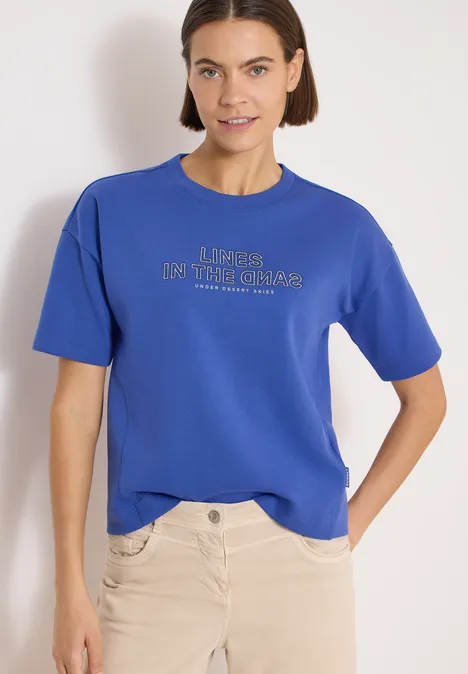 Bluza z krótkim rękawem z haftem deep river blue Osoba na jasnym tle nosi niebieski t-shirt z napisem i jasne spodnie.