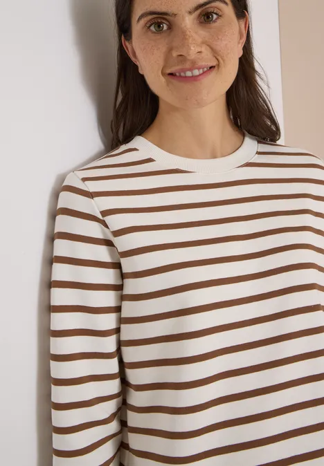 3/4-Arm Sweatshirt mit Rundhals und Streifen creamy mocha Frau lehnt an weißer Wand, trägt weiß-braun gestreiften Pullover und lächelt leicht.