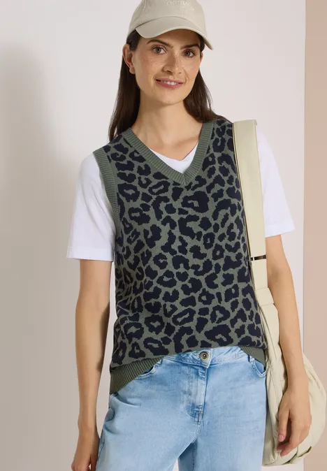 Frau in Schirmmütze trägt leo-print Weste über weißem Shirt und Jeans, mit Schultertasche, lächelt.