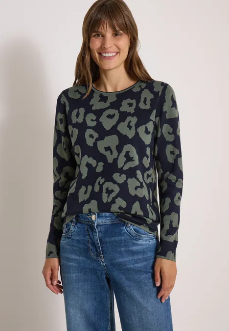 Vrouw lacht in casual trui met animalprint en blauwe jeans, staande voor een lichte achtergrond.
