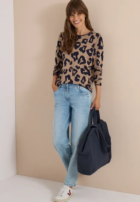 Vrouw tegen muur in pantertrui, jeans, witte sneakers en grote marineblauwe tas, ze lacht.