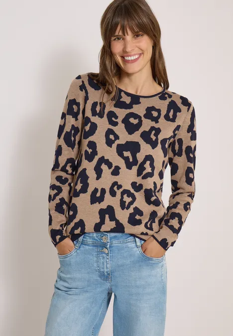 Vrouw lacht in beige zwart gevlekt trui en jeans tegen lichte achtergrond.