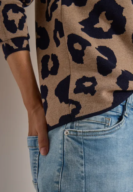Persoon in beige-blauwe trui met luipaardprint en lichtblauwe spijkerbroek, hand in zak