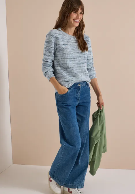 Femme souriante en jean large et pull gris-bleu à motifs, tenant une veste verte, devant un fond beige.